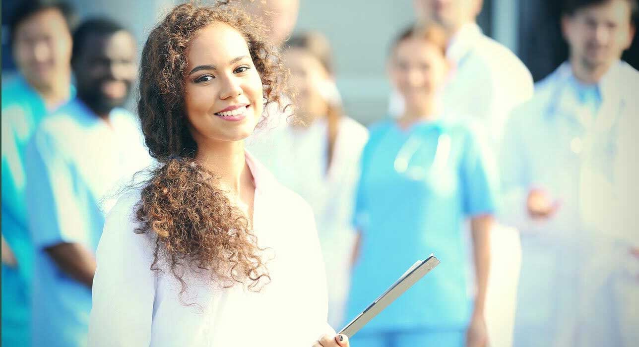 A woman with curly hair and a white coat stands smiling in the foreground, holding a clipboard. In the blurred background, a group of medical professionals, wearing blue scrubs and white coats, are gathered together.