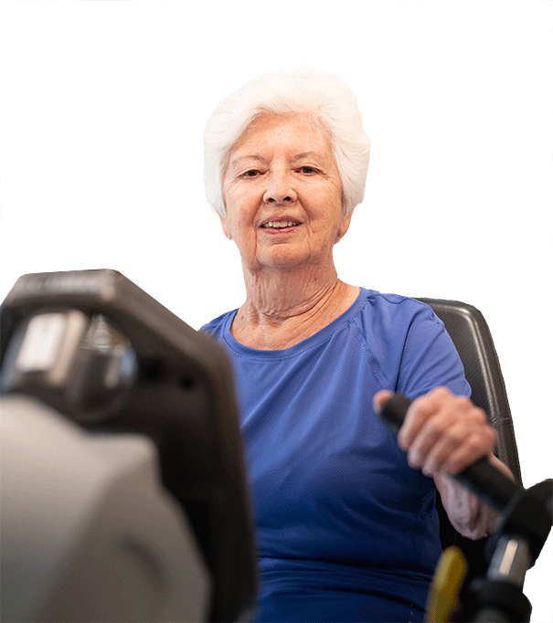 An elderly woman with white hair and a blue shirt is smiling while using exercise equipment, holding the handles and looking forward.