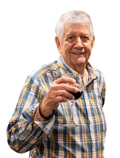 An older man with gray hair, dressed in a plaid shirt, smiles while holding a glass of red wine and raising it in a toast—celebrating the joys of Independent Living.