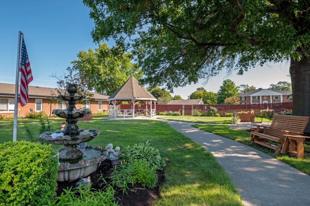 A serene garden features a fountain, a flagpole with an American flag, lush greenery, and a winding pathway leading to a gazebo. A wooden bench sits under a large tree, providing a shaded spot for relaxation. Buildings and a red fence are visible in the background.