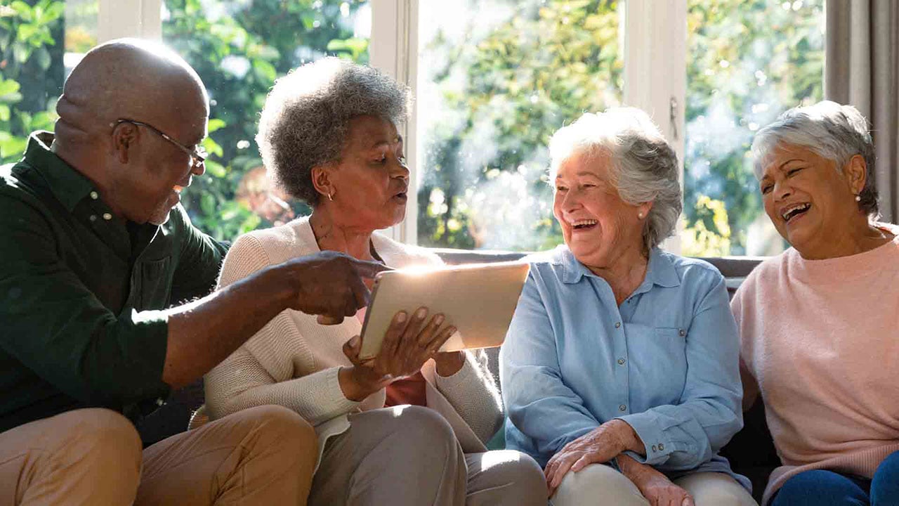 Four older adults sit on a couch, laughing and smiling together as one of them holds and points to a tablet. Sunlight streams through large windows in the background, creating a warm and cheerful atmosphere.