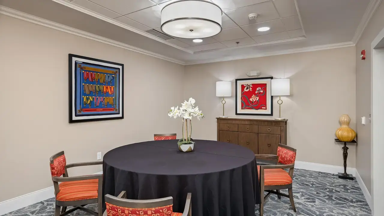 A small, well-lit conference room with a round table covered in a black tablecloth. Four chairs with red patterned cushions surround the table. On the table is a vase with white flowers. The walls have two colorful framed artworks, and a wooden sideboard stands against the back wall with two lamps.