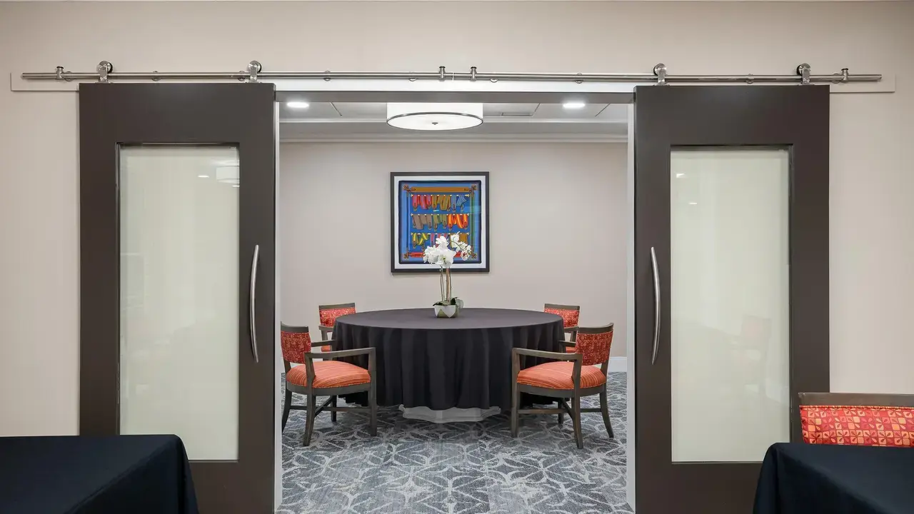 A conference room with a round table covered by a black tablecloth and surrounded by six red upholstered chairs. The room is visible through half-open sliding doors, with an artwork on the wall behind the table and a ceiling light above.