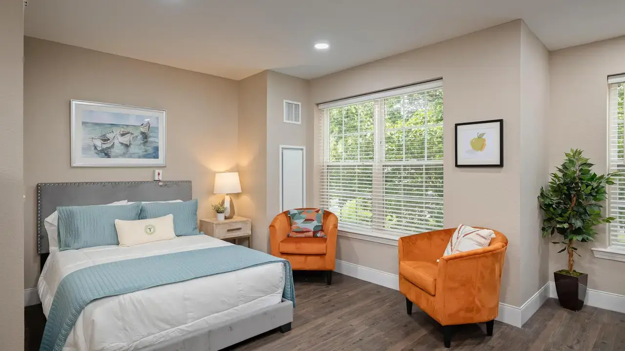 A cozy bedroom with a neatly made bed featuring blue and white bedding. A bedside table with a lamp, two orange armchairs, a small plant, framed artwork, and large windows with white blinds complete the room. The floor is dark wood, and the walls are light beige.